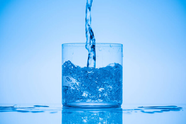 toned image of water pouring in drinking glass on blue background with splashes