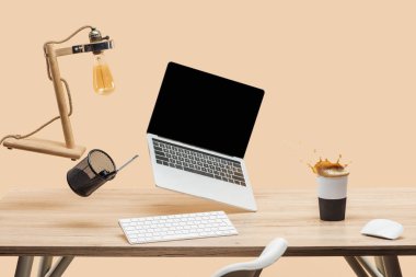 laptop with blank screen and lamp levitating in air above wooden desk with thermomug with coffee splash isolated on beige clipart
