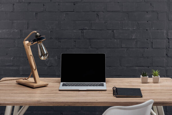 modern workplace with laptop with blank screen, decor, lamp and notebook on wooden table near black brick wall