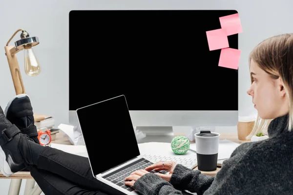 女孩坐在凌乱的工作场所 腿在桌子上 并使用在灰色隔离的笔记本电脑 — 图库照片
