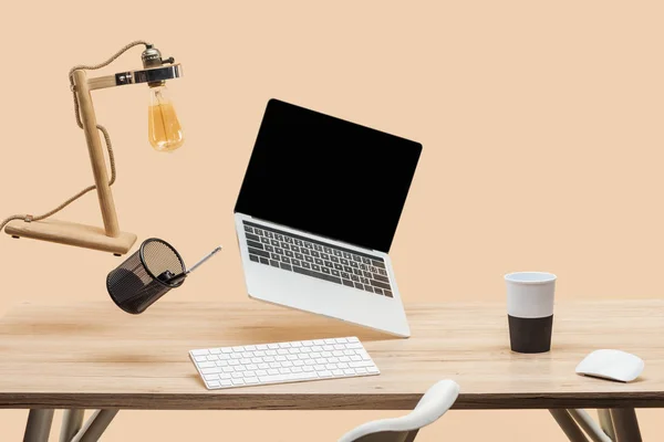 laptop with blank screen, lamp and stationery levitating in air above wooden desk isolated on beige
