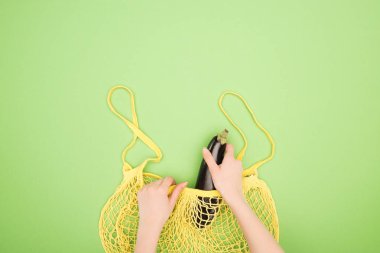cropped view of woman putting eggplant in yellow string bag on light green background clipart