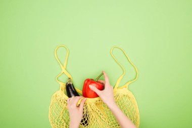 Açık yeşil arka planda sarı dize torbada kadın elleri, kırmızı biber ve patlıcan kırpılmış görünümü