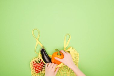partial view of woman taking fresh bell pepper out of yellow string bag with whole vegetables on light green surface clipart