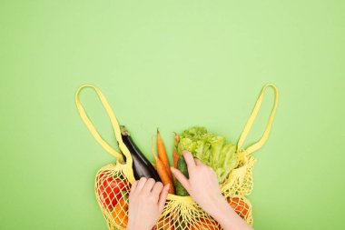 partial view of woman taking fresh cucumber out of yellow string bag with whole vegetables on light green surface clipart