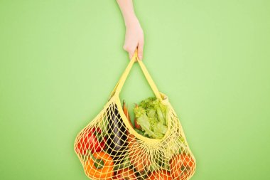 partial view of woman holding string bag with whole vegetables on light green  clipart