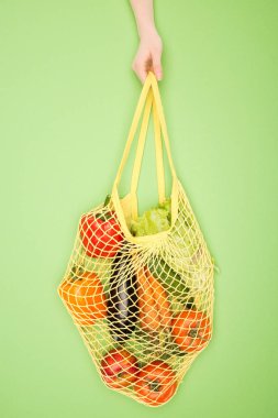 partial view of woman holding string bag with vegetables on light green  clipart