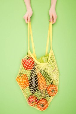 cropped view of woman holding string bag with vegetables on light green  clipart