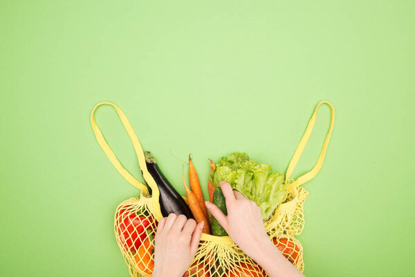 partial view of woman taking fresh cucumber out of yellow string bag with whole vegetables on light green surface