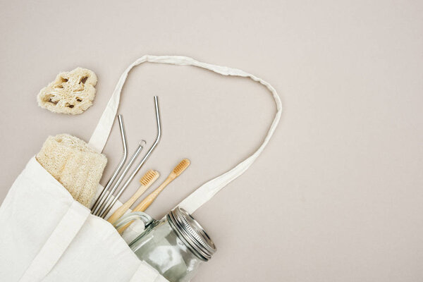 bamboo toothbrushes, organic loofah, jar and stainless steel straws in white cotton bag on grey background with copy space