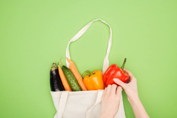 cropped view of woman putting colorful vegetables in cotton bag on light green background