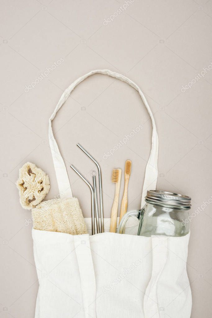 bamboo toothbrushes, organic loofah, jar and stainless steel straws in white cotton bag on grey background