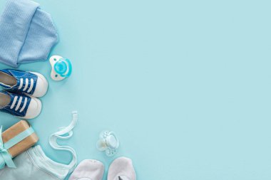 top view of pacifiers, gift, sneakers, bonnet, booties, hat on blue background with copy space clipart