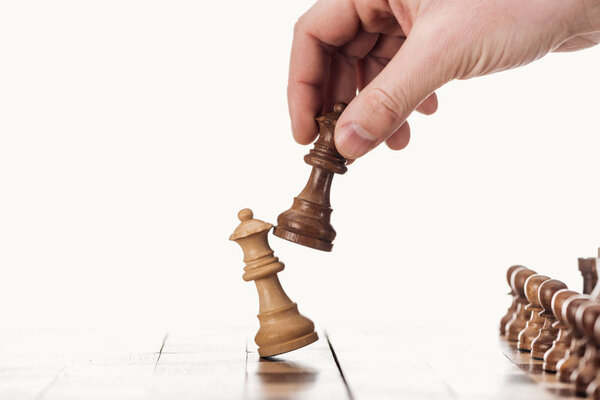 частичный вид человека, держащего коричневую королеву рядом с бежевой королевой на деревянной шахматной доске, изолированной на белом
