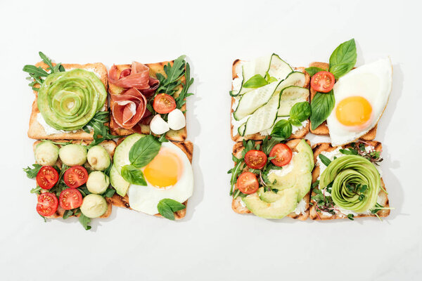 Top view of toasts with cut vegetables and prosciutto on white surface