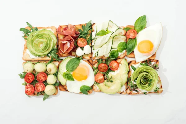Top view of toasts with cut vegetables and prosciutto on white surface