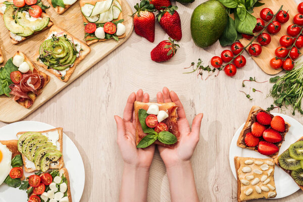 top view of woman holding toast with cherry tomatoes and prosciutto over wooden table with greenery and ingredients