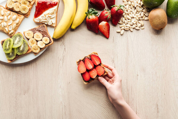 top view of woman holding fruit toast at wooden table with fresh ingredients 