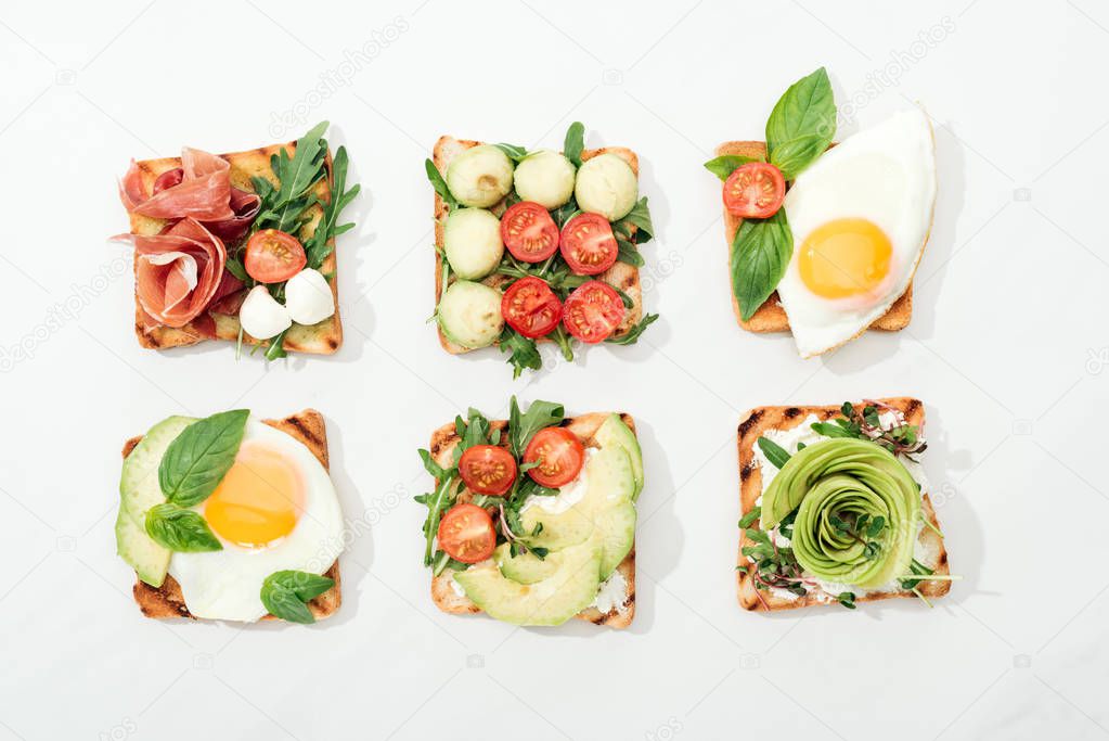Top view of toasts with cut vegetables and prosciutto on white surface