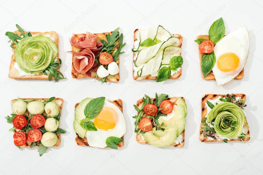Top view of toasts with cut vegetables and prosciutto on white surface