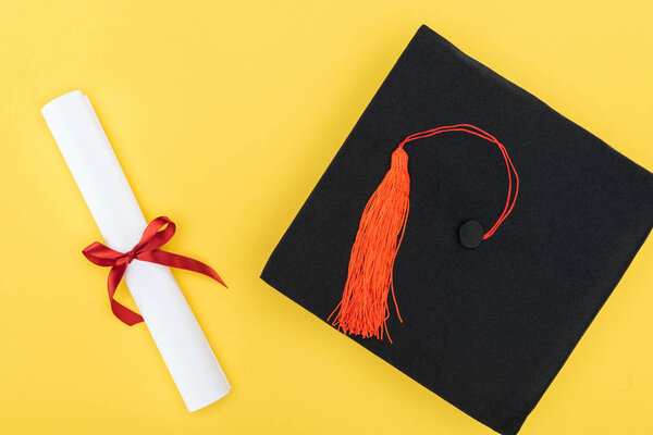 Top view of academic cap and diploma with red ribbon isolated on yellow