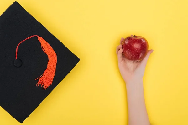 Обрізаний Вид Жінки Академічною Шапочкою Тримає Червоне Яблуко Жовтій Поверхні — стокове фото