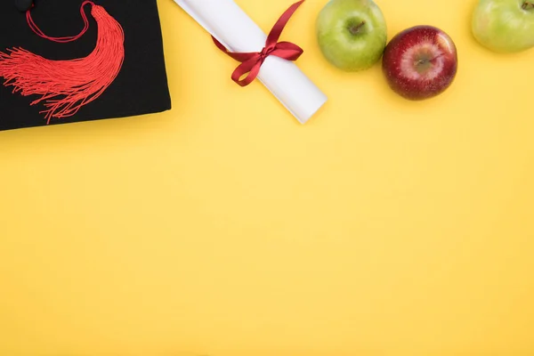 Top view of academic cap, diploma and apples on yellow surface