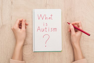 cropped view of woman holding marker near notebook with what is autism question on wooden table clipart