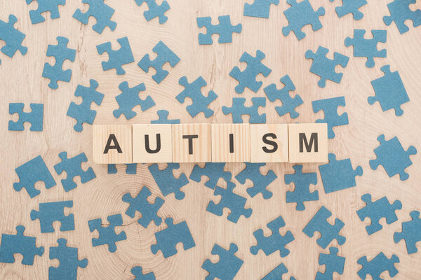 top view of autism lettering made of wooden blocks among blue puzzle pieces on wooden table