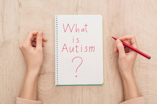 cropped view of woman holding marker near notebook with what is autism question on wooden table