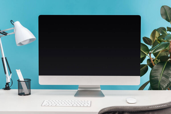 workplace with computer, lamp, stationery on white table near green plant on blue background