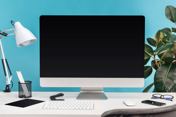 рабочее место с гаджетами, лампой, канцелярскими принадлежностями на белом столе возле зеленого завода на синем фоне
