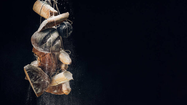 white flour falling at loaves of bread, baguettes and croissant on ropes on black background with copy space