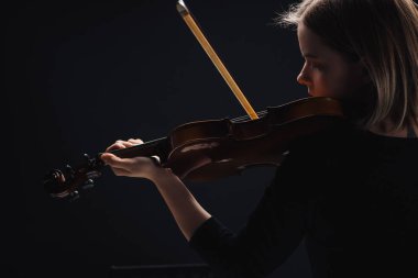 young concentrated woman playing cello with bow in darkness isolated on black  clipart