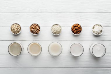 top view of glasses with coconut, chickpea, oat, rice and almond milk on white wooden surface with ingredients in bowls clipart