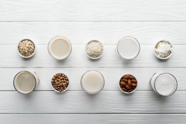 top view of glasses with coconut, chickpea, oat, rice and almond milk on white wooden table with ingredients in bowls clipart