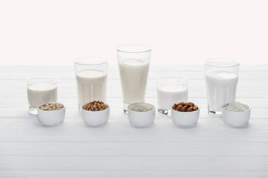 glasses with coconut, chickpea, oat, rice and almond milk with ingredients in bowls isolated on white clipart