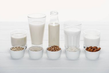 glasses and bottle with coconut, chickpea, oat, rice and almond milk on white wooden table with ingredients in bowls isolated on white clipart