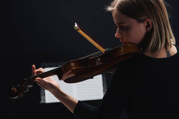 selective focus of young concentrated woman playing cello with bow in darkness with music book at background isolated on black 