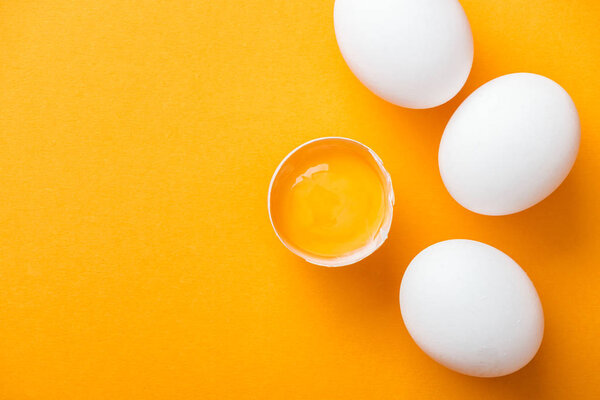 top view of smashed chicken egg with yolk on bright orange background among white whole eggs