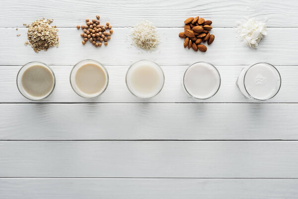 top view of glasses with coconut, chickpea, oat, rice and almond milk on white wooden surface with ingredients