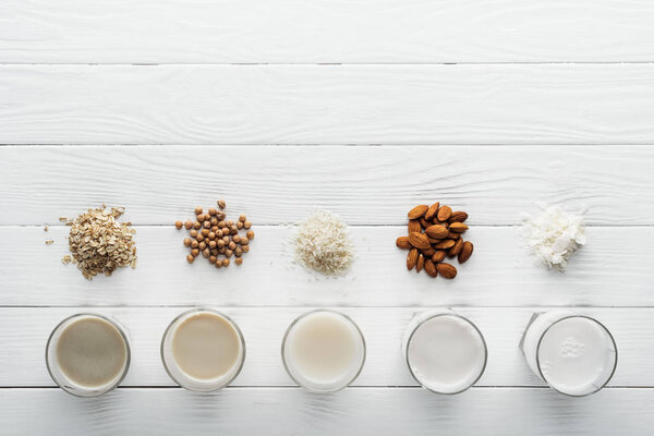 top view of glasses with coconut, chickpea, oat, rice and almond milk on white wooden table with ingredients