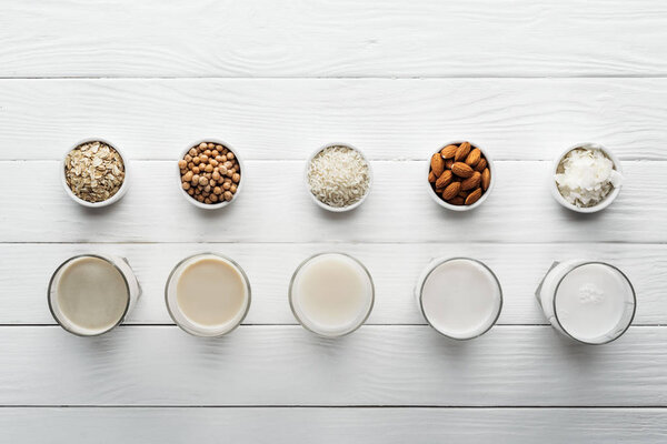 top view of glasses with coconut, chickpea, oat, rice and almond milk on white wooden surface with ingredients in bowls