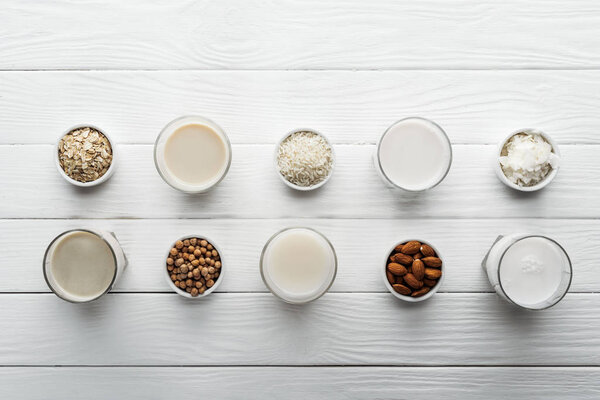 top view of glasses with coconut, chickpea, oat, rice and almond milk on white wooden table with ingredients in bowls