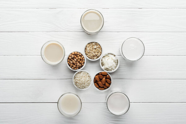 top view of glasses with coconut, chickpea, oat, rice and almond milk on white surface with ingredients in bowls