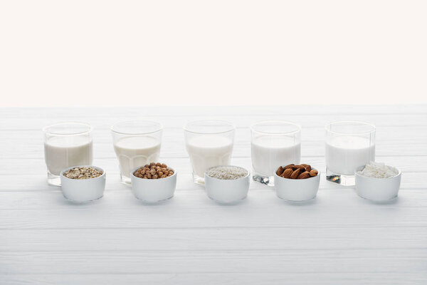 glasses with coconut, chickpea, oat, rice and almond milk on white wooden surface with ingredients in bowls isolated on white