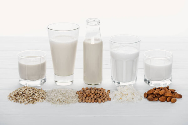 glasses and bottle with coconut, chickpea, oat, rice and almond milk on white wooden surface with ingredients isolated on white