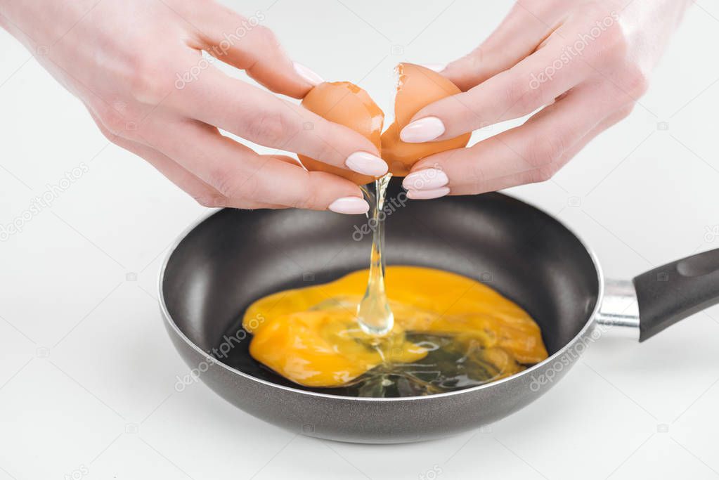 cropped view of woman smashing egg while preparing scrambled eggs in pan on white background