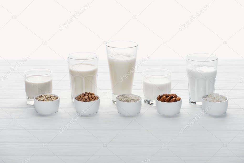 glasses with coconut, chickpea, oat, rice and almond milk with ingredients in bowls isolated on white