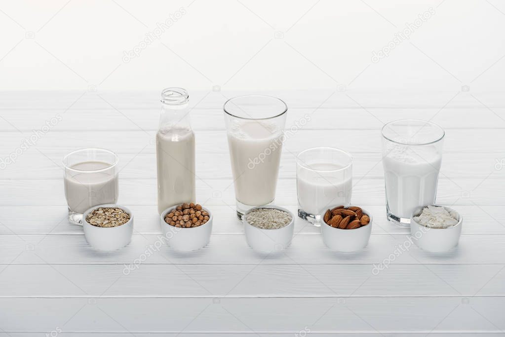 glasses and bottle with coconut, chickpea, oat, rice and almond milk on white wooden surface with ingredients in bowls isolated on white
